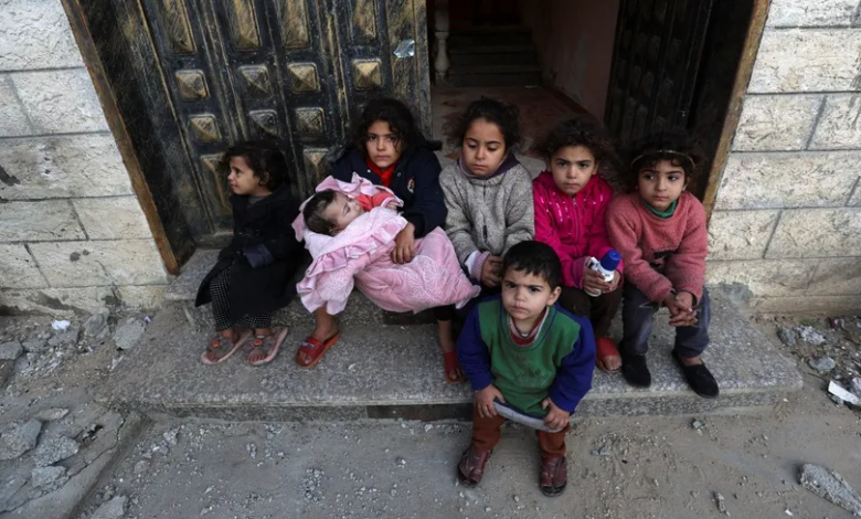 طفلة بين أطفال آخرين تحمل شقيقها الرضيع أمام منزلهم في قطاع غزة بينما دمرت الغارات الإسرائيلية عدداً من المنازل المحيطة. 17 ديسمبر 2023