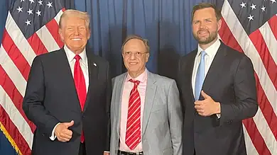 Donald Trump with two men at a political event, posing in front of American flags.